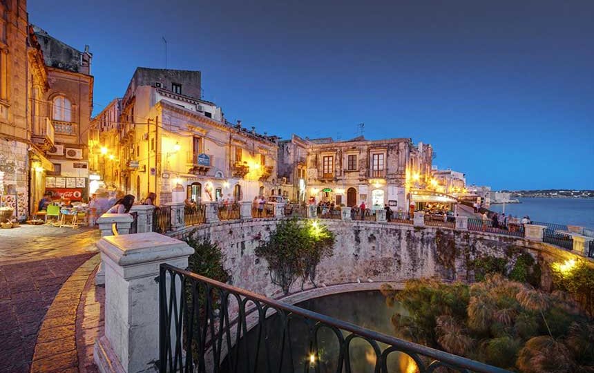 Vista di un affascinante angolo di città con edifici storici illuminati, un ponte che conduce a una piazza, e un laghetto riflettente al crepuscolo.